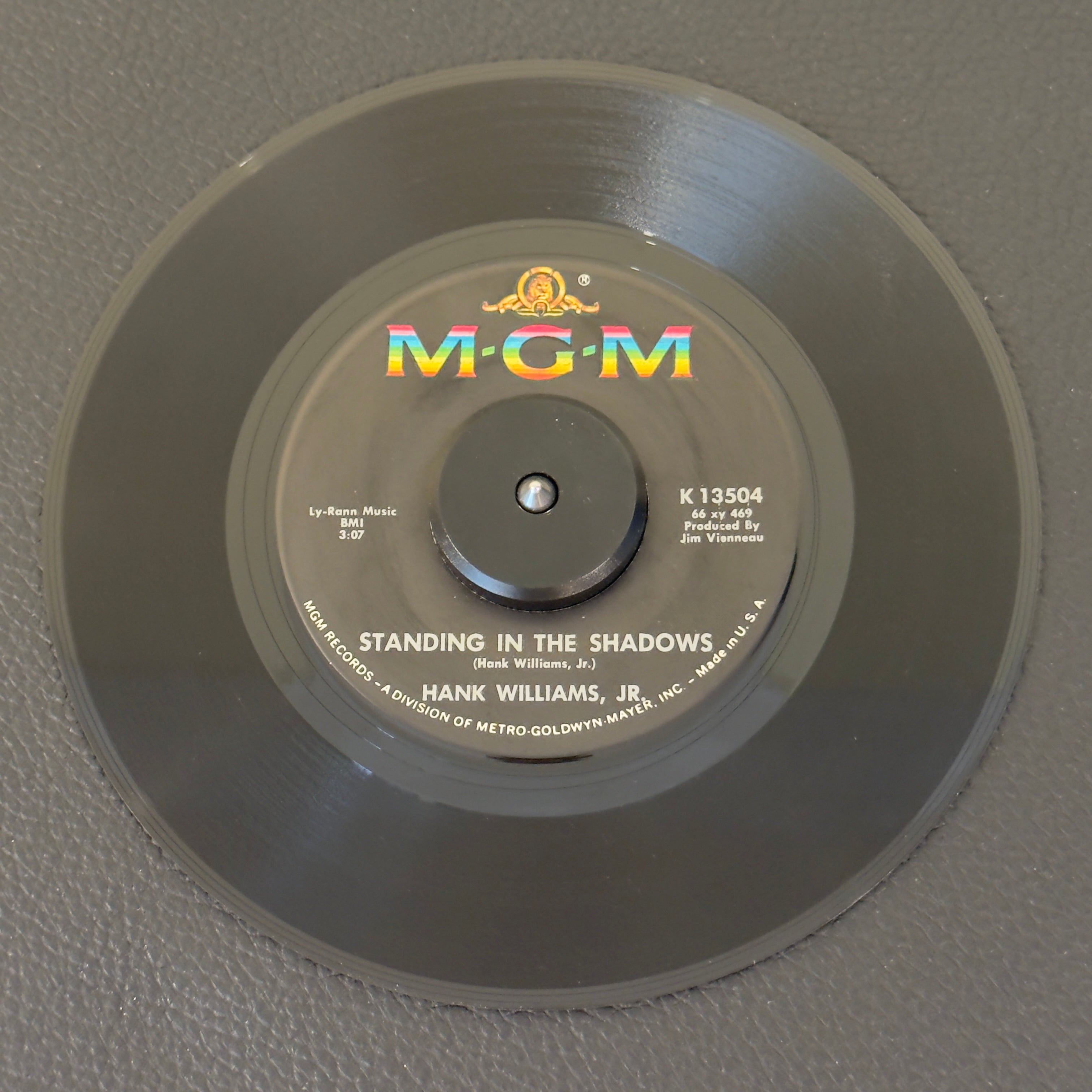 7" vinyl record by Hank Williams Jr. featuring "Standing In The Shadows/It's Written All Over Your Face", released on MGM Records (K13504), in VG+ (Very Good Plus) condition, classic analog collectable for turntable playback. Original MGM sleeve included.
